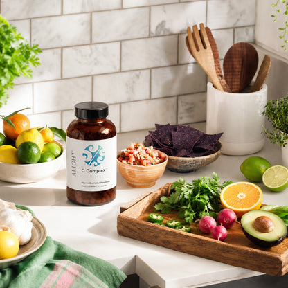 A bottle of Aight C Complex dietary supplement placed on a kitchen counter surrounded by fresh fruits, vegetables, and wooden utensils.