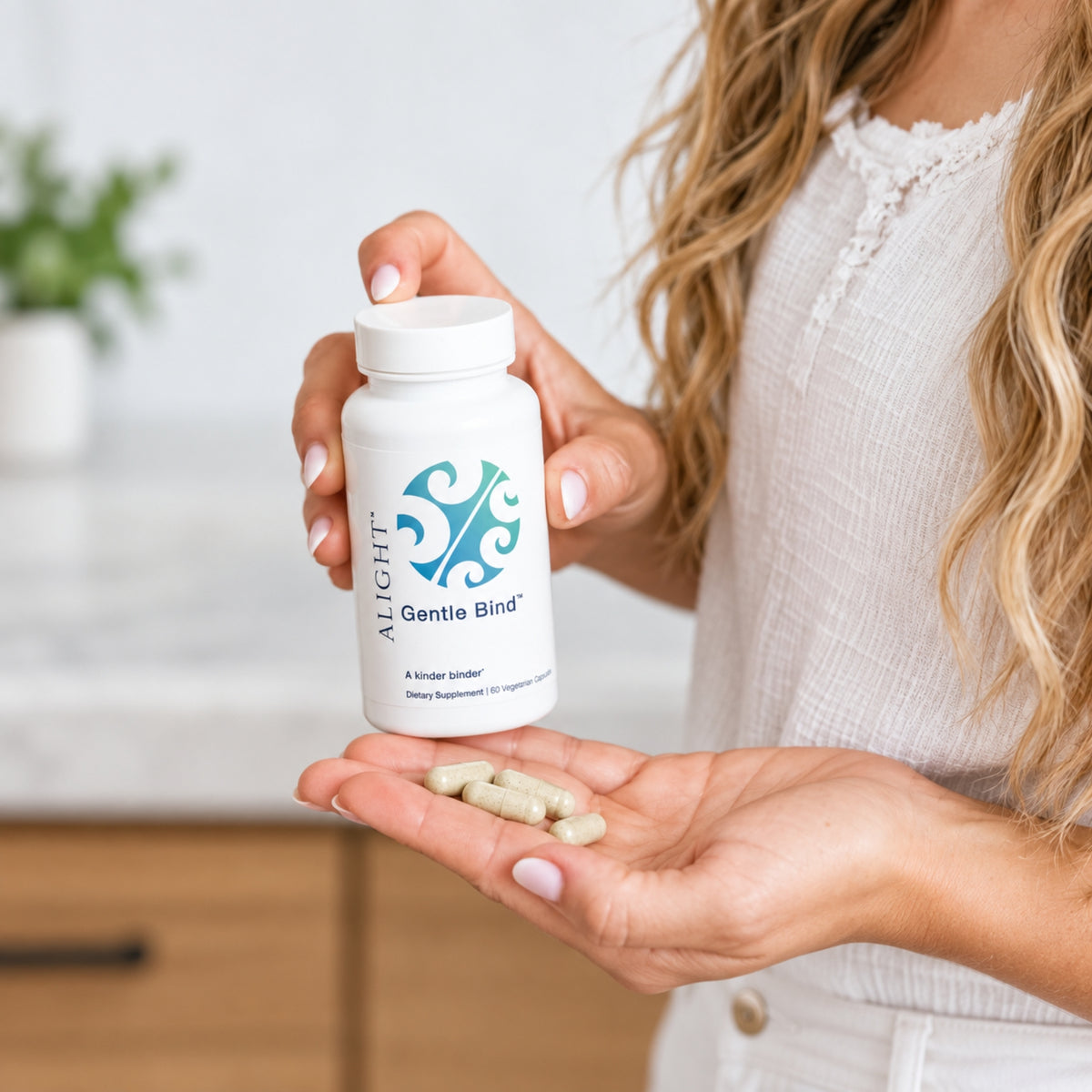 A woman holds a white bottle labeled  Gentle Bind dietary supplement in one hand while holding several beige capsules in the other, set against a softly blurred kitchen background.