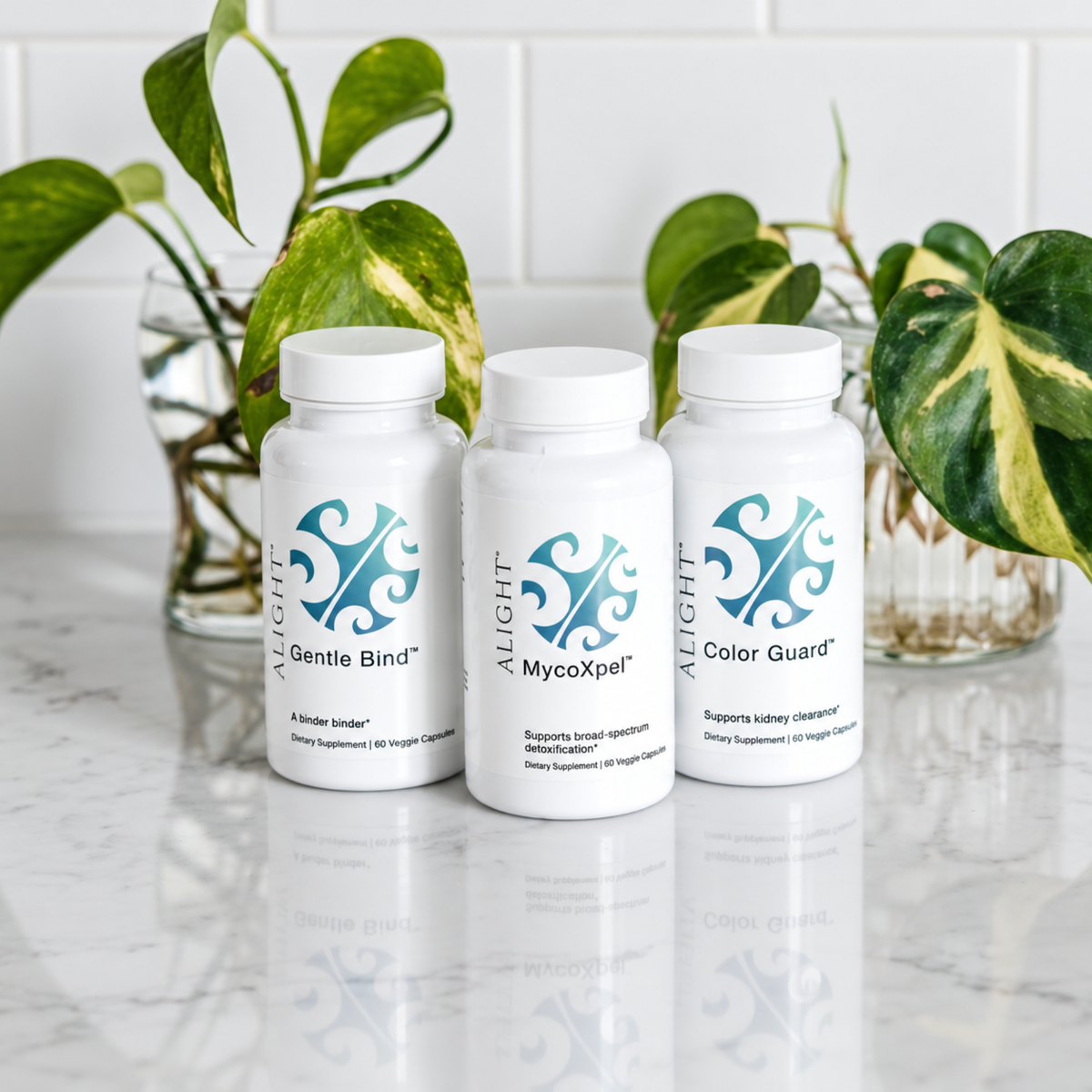 Three white supplement bottles from the brand ALiGHT arranged on a reflective white surface with green houseplants in the background. The bottles are labeled Gentle Bind, MycoXpel, and Color Guard.