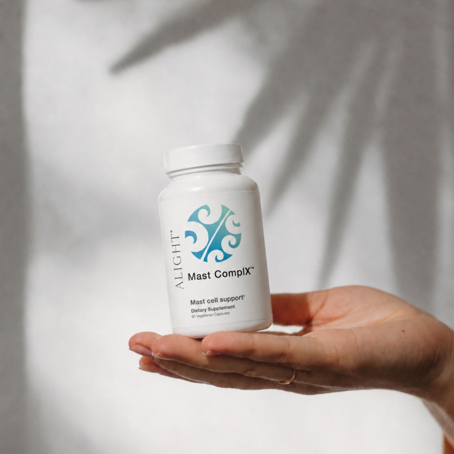 A hand holding a white bottle of Alight Mast Compix Mast cell support dietary supplement against a soft, shadowed light gray background.