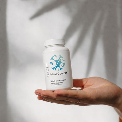 A hand holding a white bottle of Alight Mast Compix Mast cell support dietary supplement against a soft, shadowed light gray background.